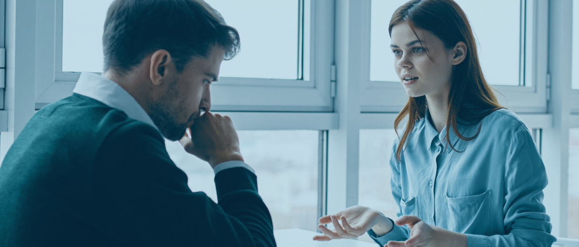 office workplace with man in jumper and shirt looking down while woman in blue shirt holds out hands to plead and explain